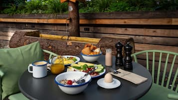 Una mesa redonda con alimentos para el desayuno, como cuencos de cereal, tostadas y huevos, colocada al aire libre con sillas verdes y un banco de madera al fondo.