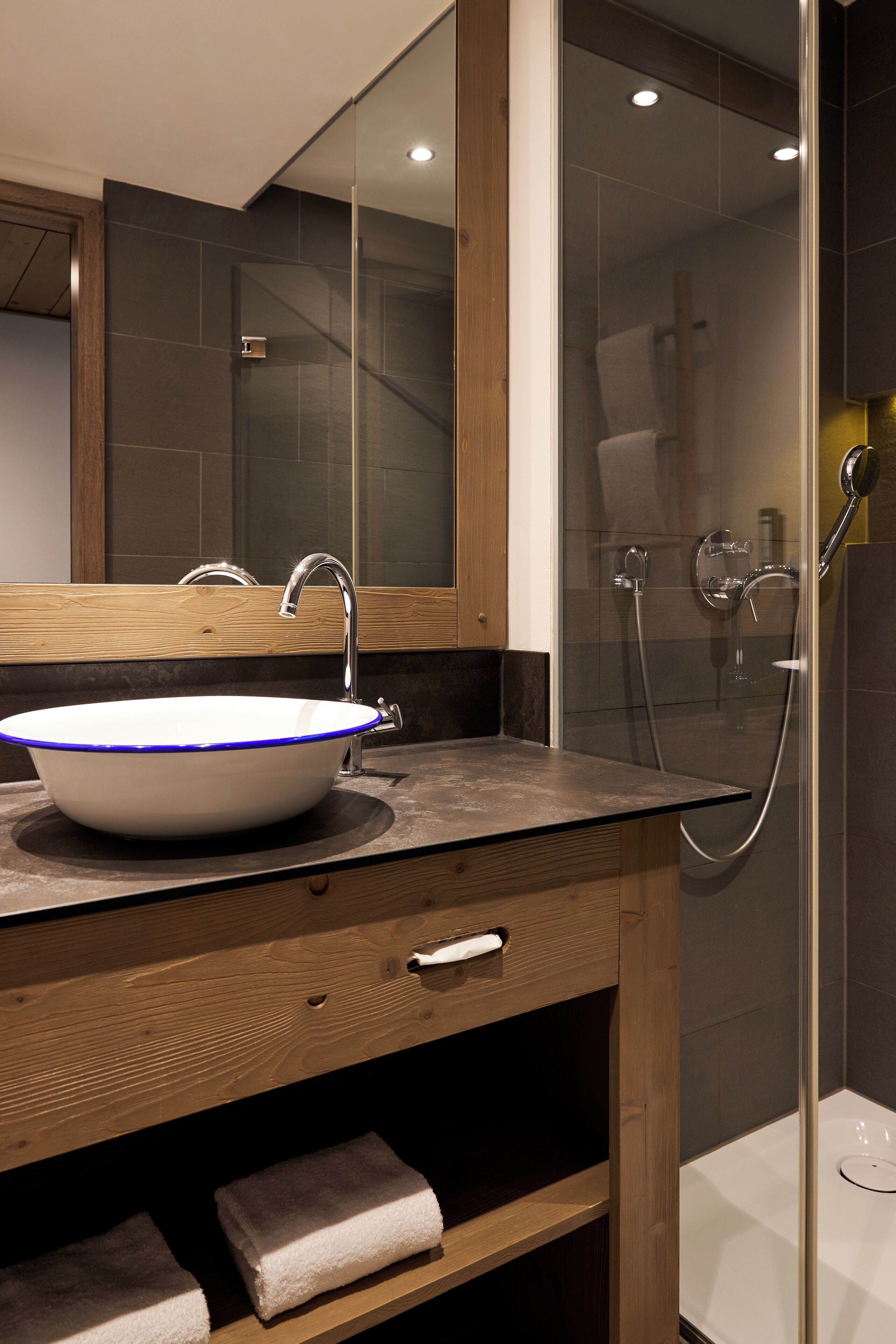 Ein modernes Badezimmer mit einem großen Spiegel, einem weißen Waschbecken mit blauen Akzenten, einer Holz-Kommode und einer Duschkabine aus Glas.