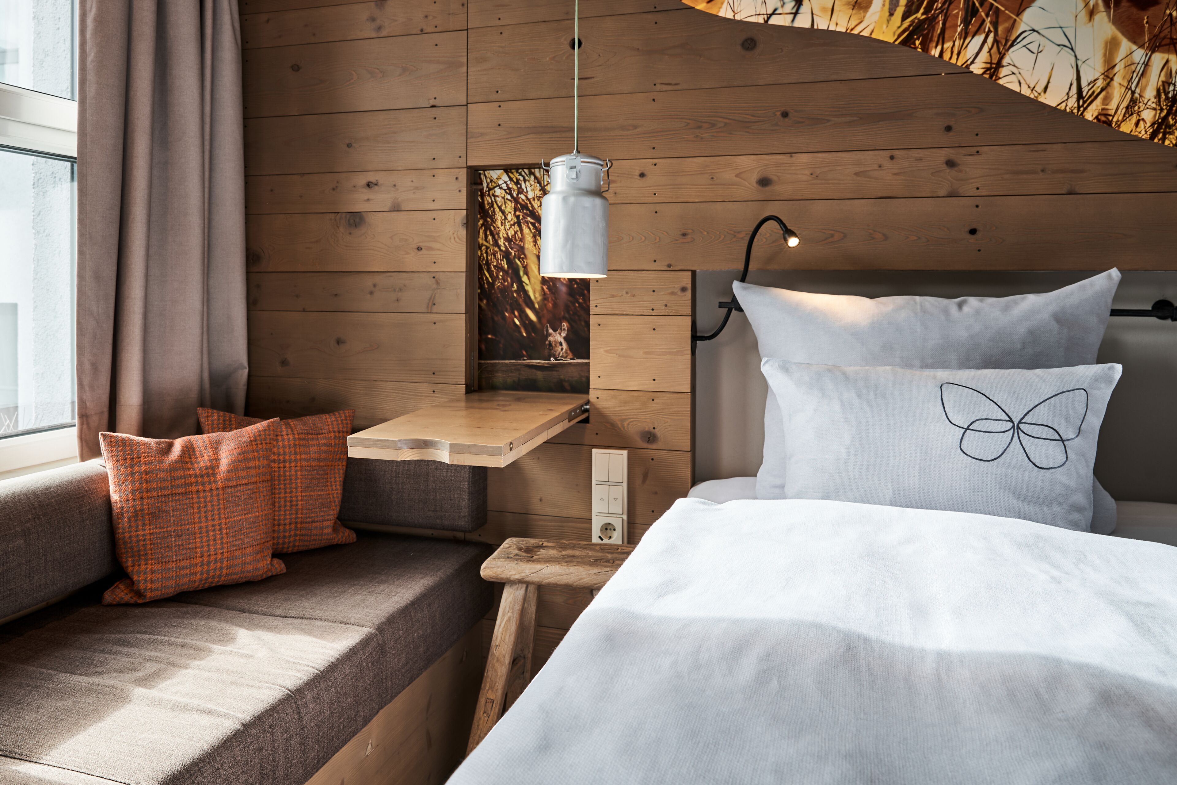 Een gezellige slaapkamer met een houten hoofdbord, een nachtkastje, een raam met gordijnen, en een bed met witte beddengoed en een kussen met vlinderprint.