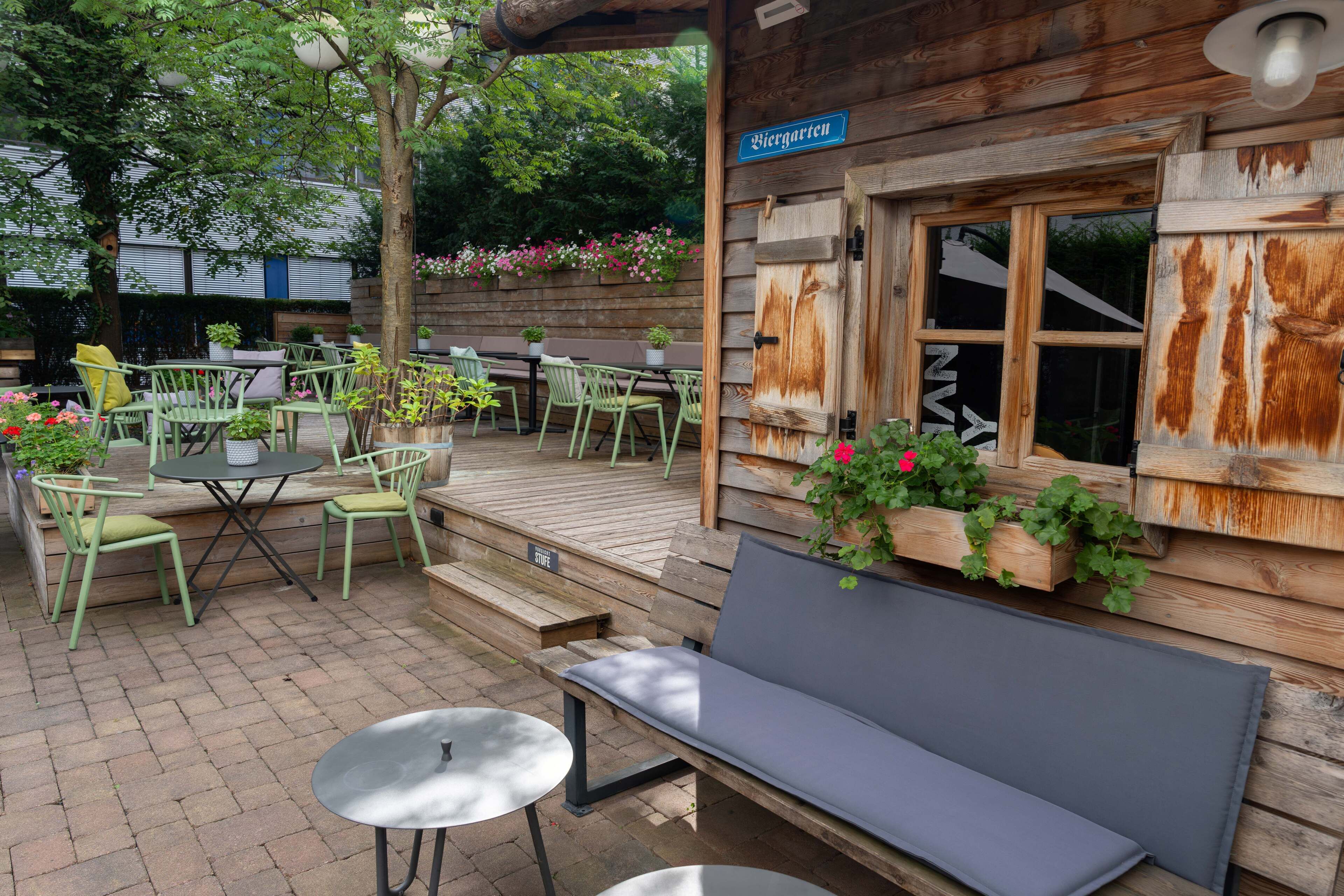 A wooden outdoor seating area with green chairs and tables, a bench, and flower boxes on the wall.