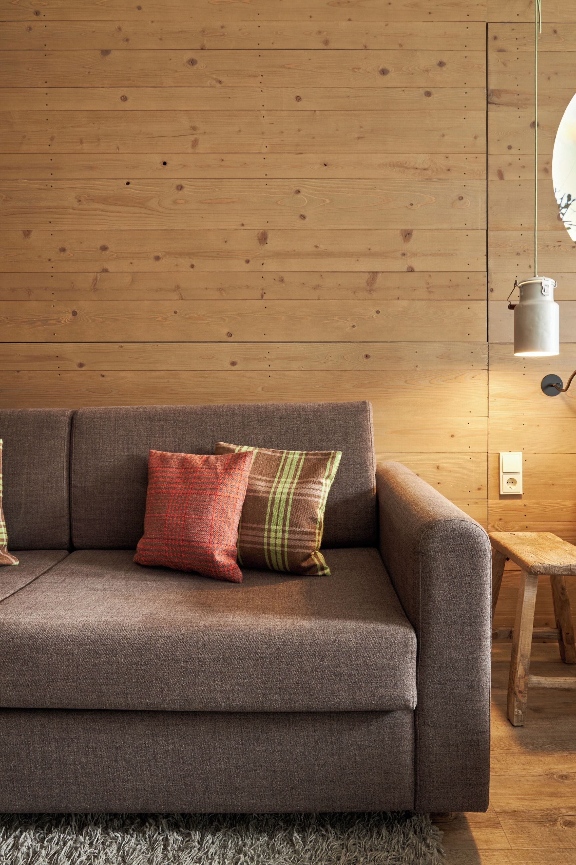 A wooden-paneled room with a grey sofa, two pillows, a small wooden stool, and a hanging light fixture.