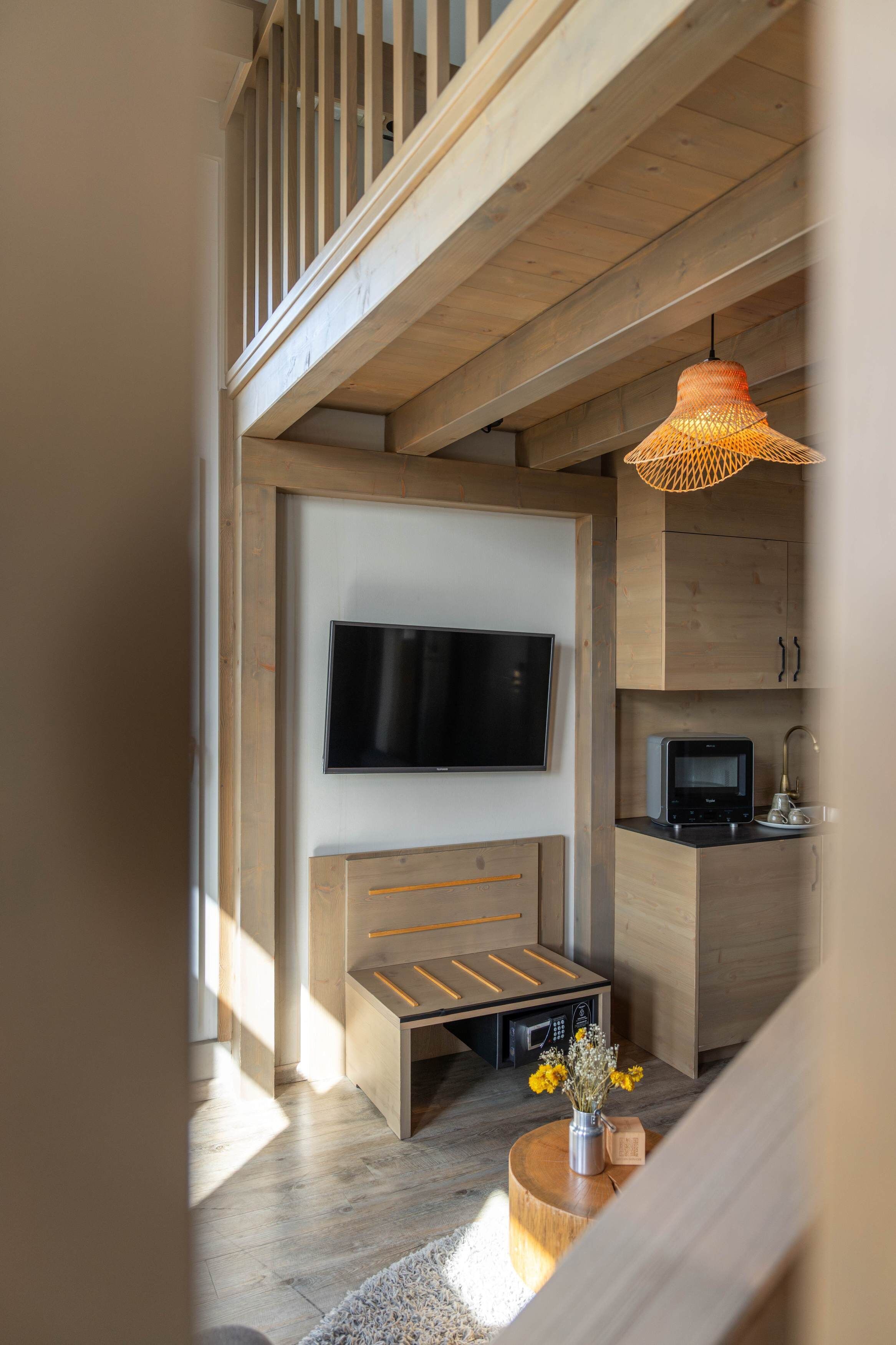 A modern living space with a wooden staircase, a flat-screen TV mounted on the wall, a built-in bench with drawers, a small kitchen area with a microwave, and a round wooden coffee table with a vase of flowers.