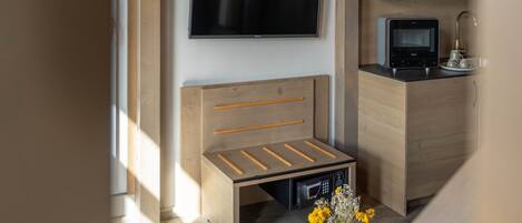 A modern living space with a wooden staircase, a flat-screen TV mounted on the wall, a built-in bench with drawers, a small kitchen area with a microwave, and a round wooden coffee table with a vase of flowers.