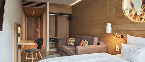 A modern hotel room with a wooden wall, a bed with white bedding, a sofa with colorful pillows, a desk with a chair, and a wall-mounted lamp.