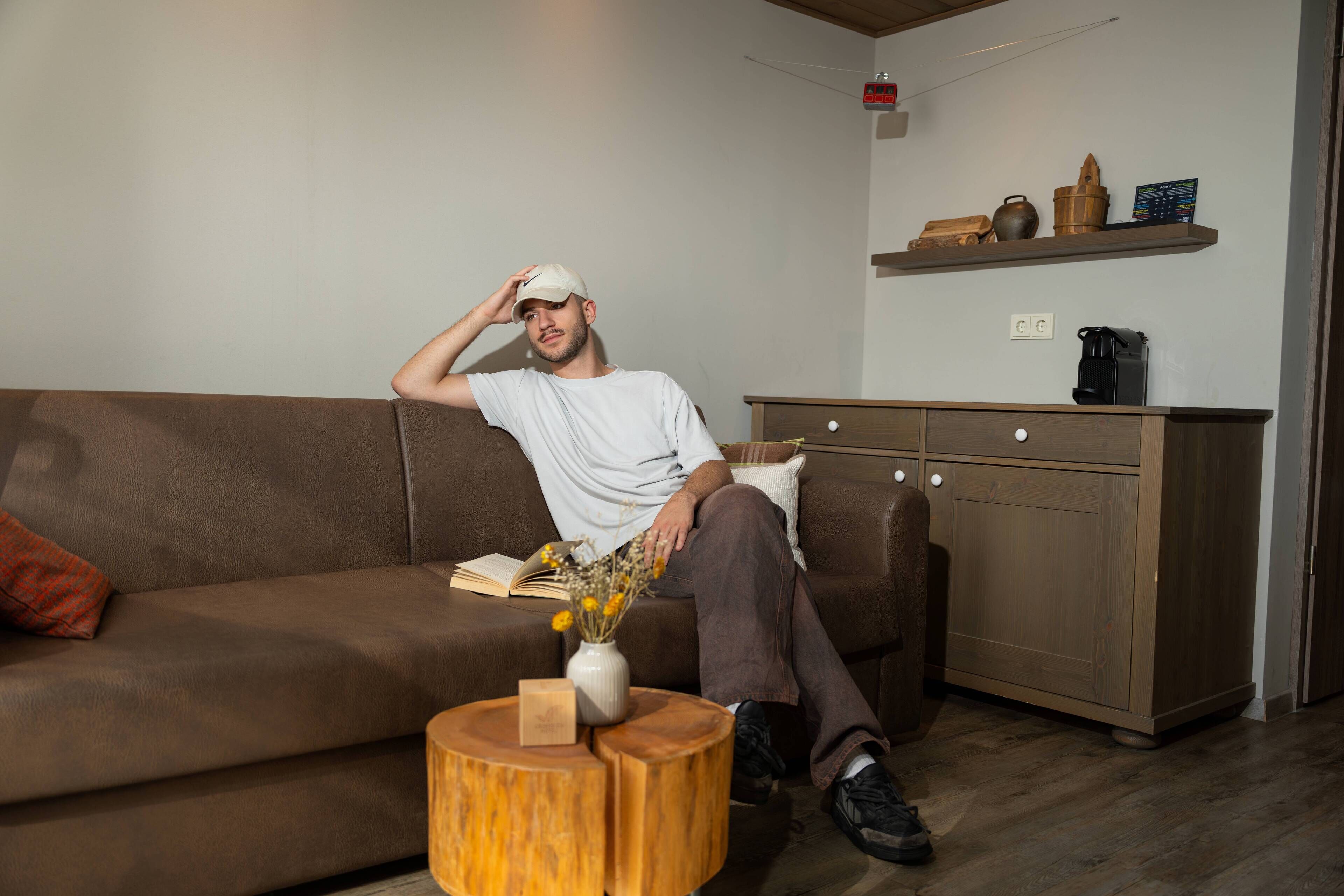 Una persona sentada en un sofá marrón en una habitación con un mueble de madera y una estantería con objetos decorativos.