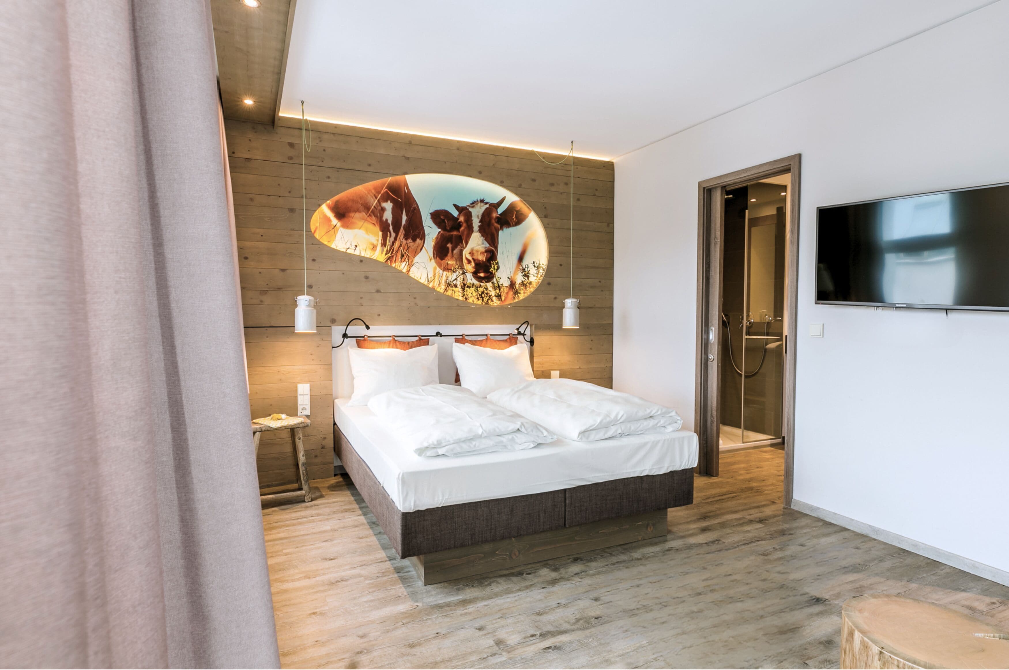 Une chambre moderne avec une tête de lit en bois, un lit recouvert de linge de maison blanc, un téléviseur fixé au mur et une œuvre d’art originale qui décore le mur.