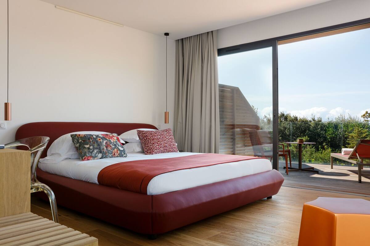 Une chambre d’hôtel moderne dotée d’un grand lit, d’un plancher en bois, d’un balcon avec coin salon et offrant une vue sur le panorama urbain.