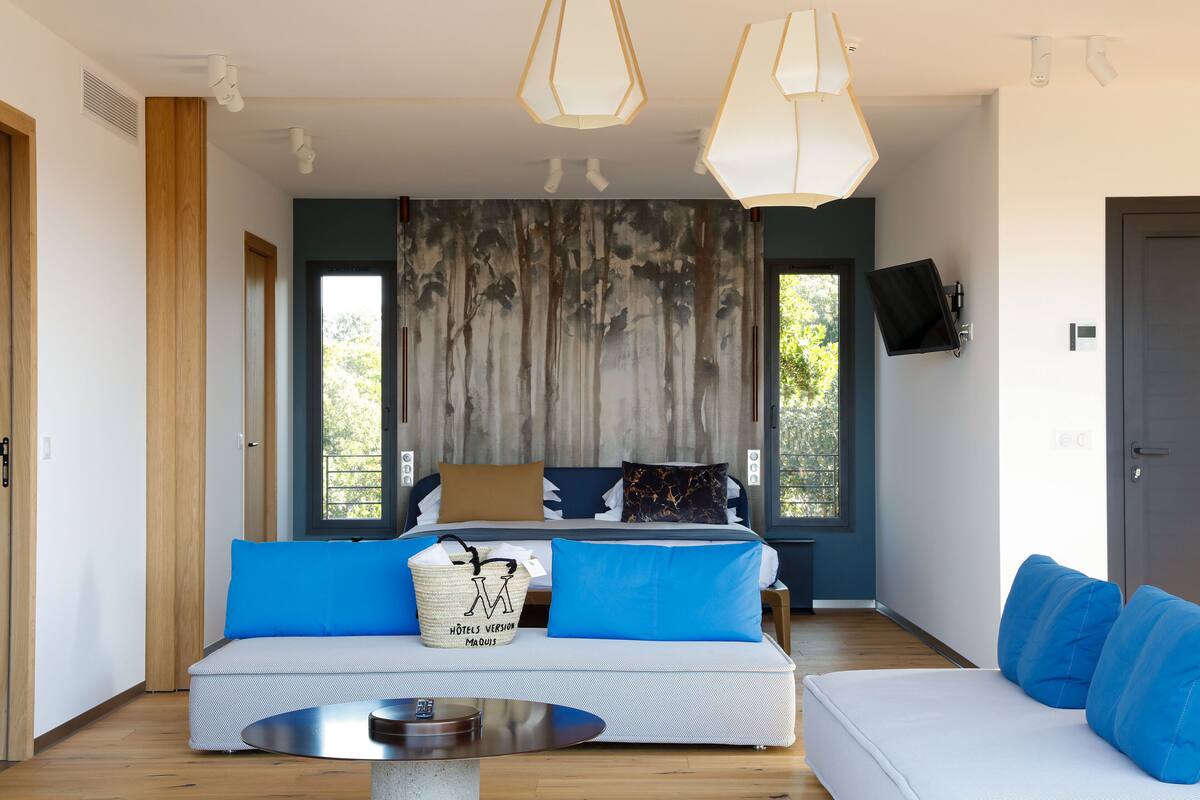 Une chambre d’hôtel moderne avec un grand lit, un canapé et une table à manger. Elle est dotée de grandes fenêtres, d’un téléviseur et d’une porte.