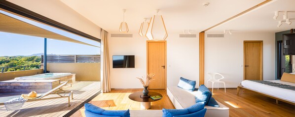 A modern living room with a sofa, a coffee table, and a TV. There is a balcony with a hot tub and a view of the landscape.