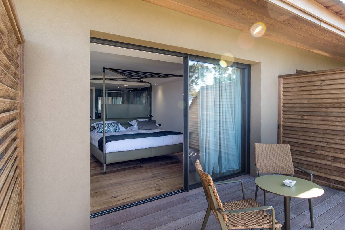 Une chambre avec un lit et une porte-fenêtre coulissante donnant sur un balcon équipé d’une table et de chaises.