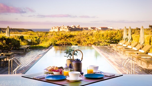 Une table de petit-déjeuner avec des assiettes, des tasses et une théière, donnant sur une piscine et une villa.