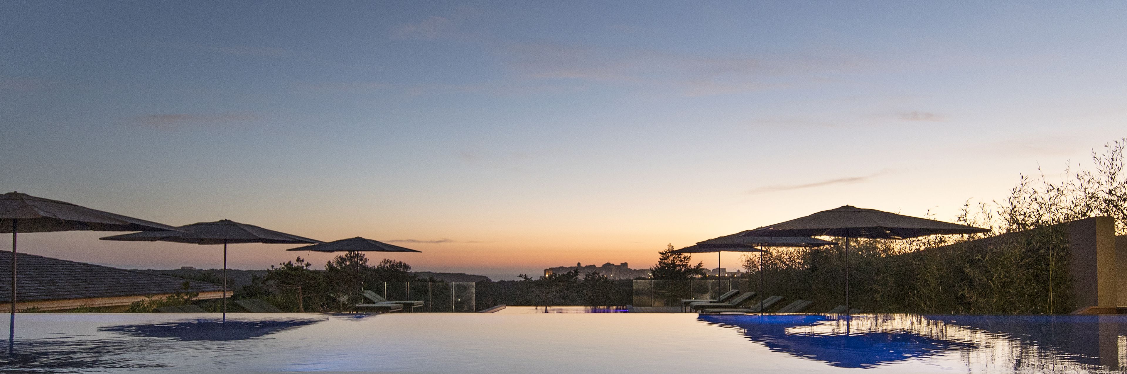 A pool area with umbrellas and lounge chairs, surrounded by trees and a city skyline at sunset.