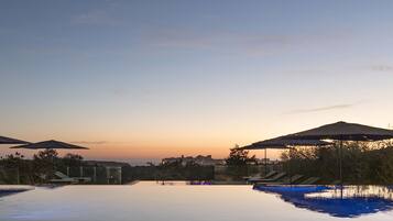A pool area with umbrellas and lounge chairs, surrounded by trees and a city skyline at sunset.
