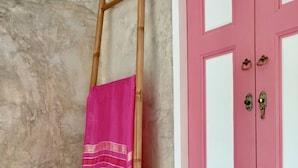 Una habitación con pared de concreto, una puerta rosa con ojo de cerradura y una escalera de madera apoyada en la pared, con una toalla rosa colgada.