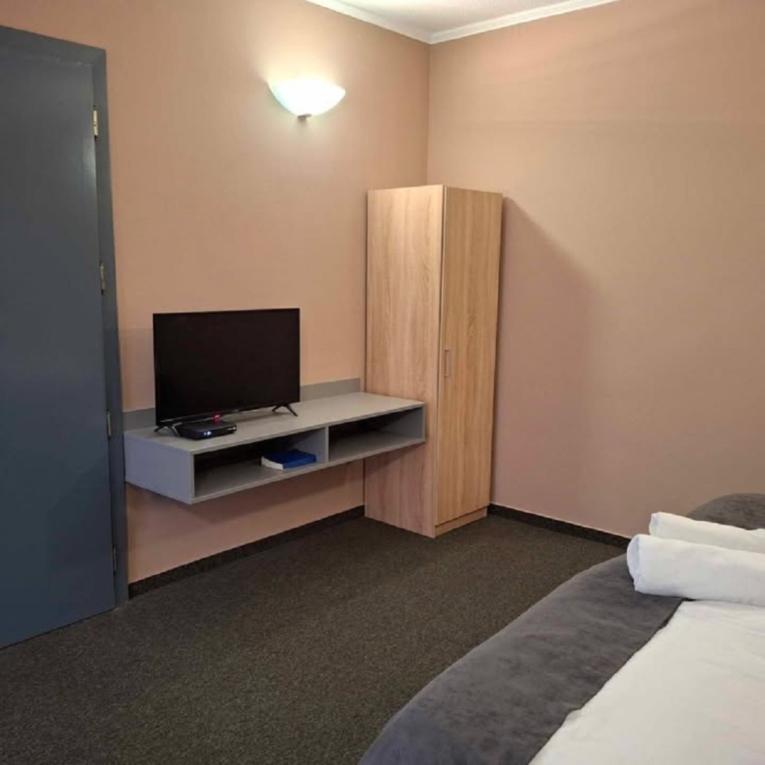 A hotel room with a bed, a TV on a stand, a wardrobe, and a wall-mounted light.