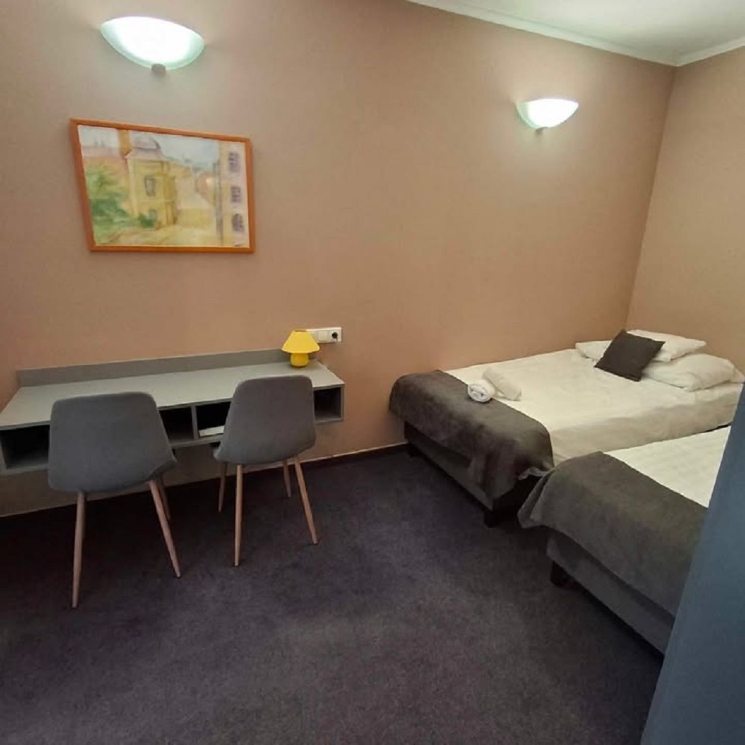 A small, neatly arranged room with a desk, two chairs, a bed, and a painting on the wall.