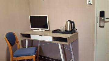 A compact hotel room with a wooden desk, a single chair, a television, a kettle, and a door with a lock.