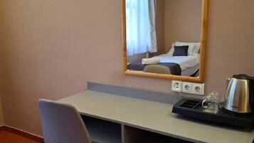 A hotel room with a desk, chair, and a bed with a white and blue bedspread. There is a mirror above the desk, and a kettle on the desk.
