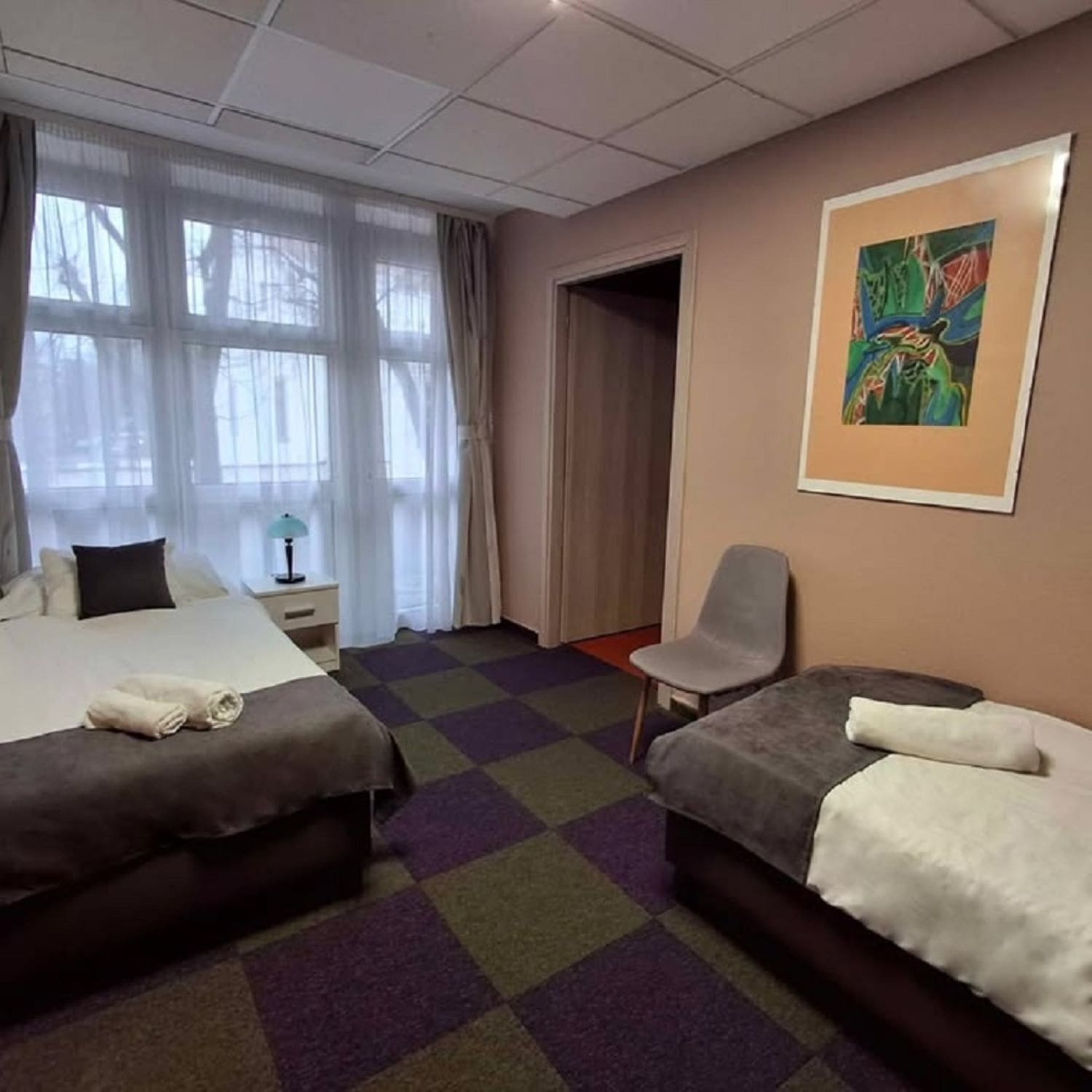 A hotel room with two beds, a chair, a painting, and a window with curtains.