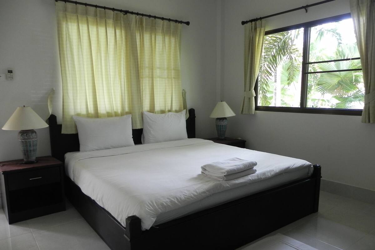 A bedroom with a bed, two nightstands, a window with curtains, and a view of greenery outside.