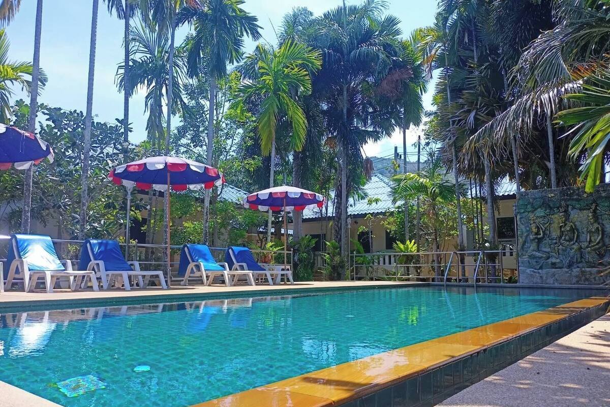 A swimming pool with clear water, surrounded by lounge chairs and umbrellas, with palm trees and a building in the background.