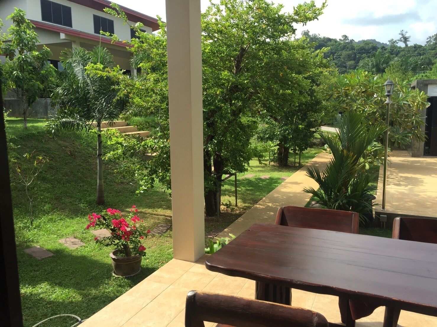 A covered outdoor patio with a wooden table and chairs, surrounded by lush greenery and a pathway leading to a building.