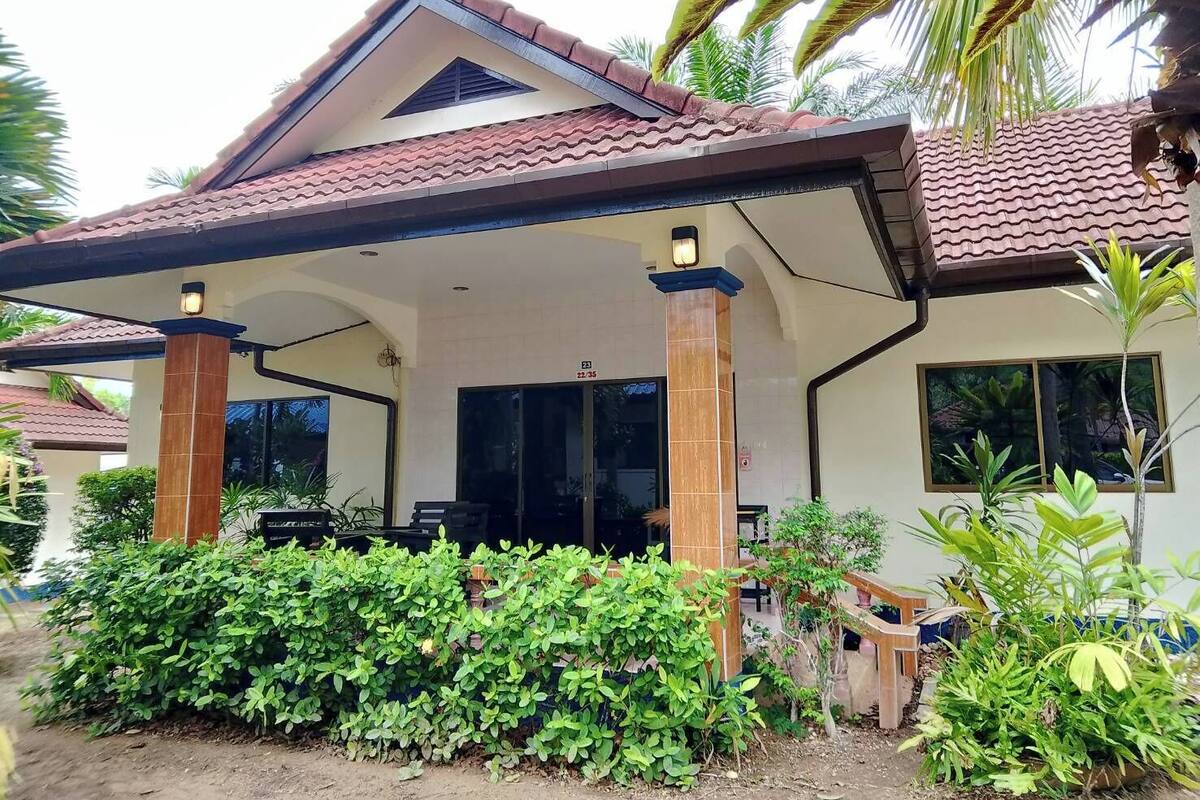 A two-story building with a tiled roof, a covered porch with a bench, and a small garden area with various plants.