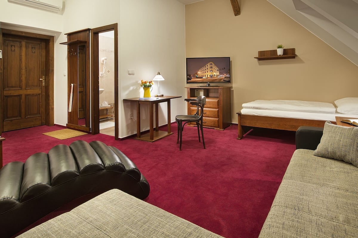 A hotel room with a bed, a sofa, a chair, a small table with a lamp, a mirror, and a painting on the wall.