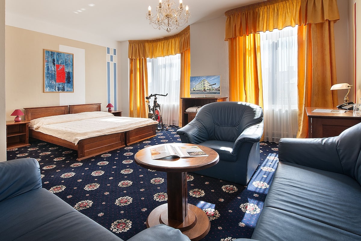 A hotel room with a bed, a sofa, a chair, a small table, and a desk with a lamp.