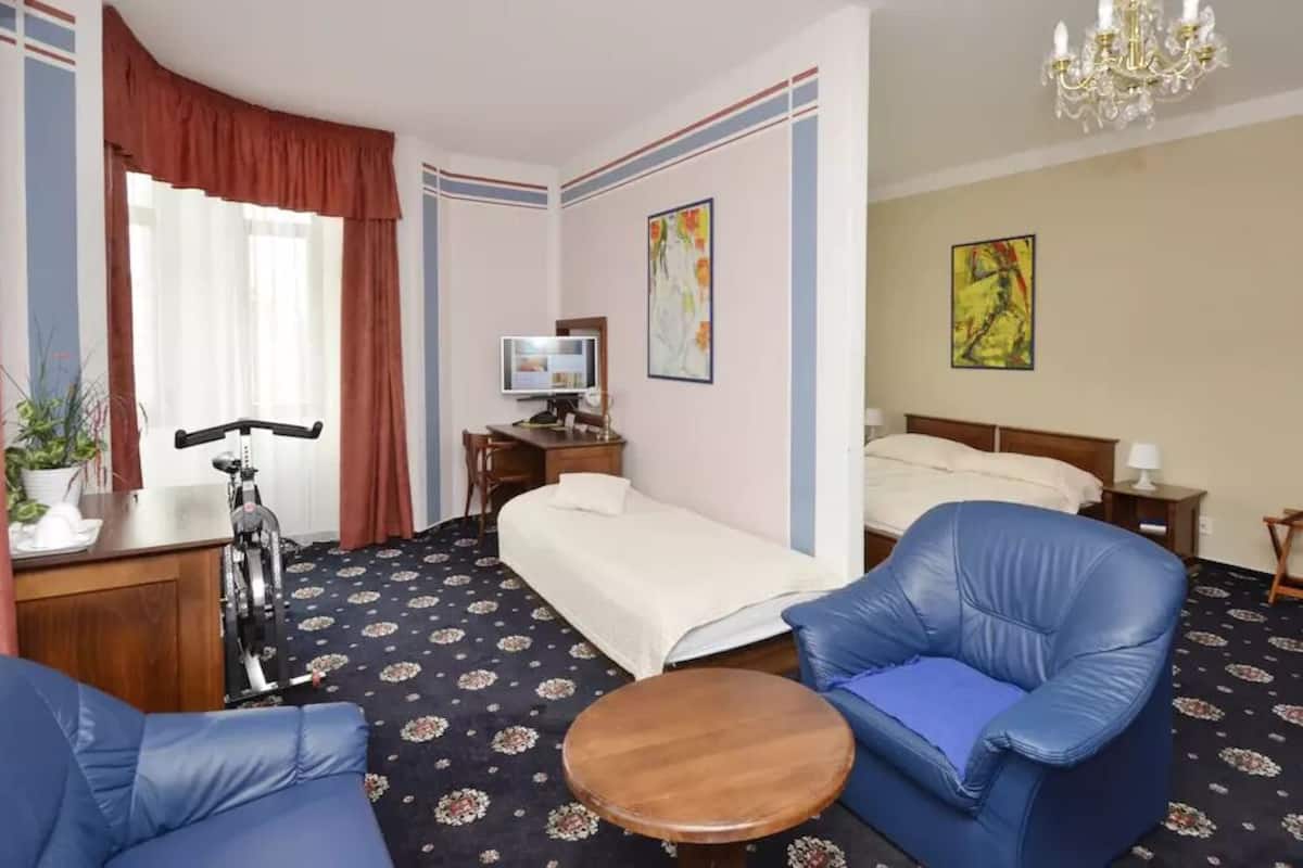 A hotel room with a bed, two blue armchairs, a small round table, a desk with a computer, a TV, and a potted plant.