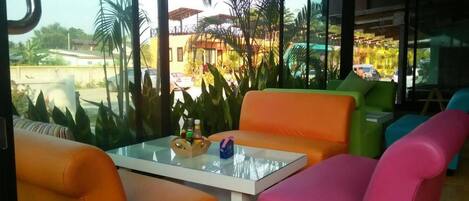 Un interior moderno con asientos naranja y rosa, una mesa de vidrio y vistas a palmeras y un edificio.