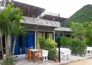 A building with a patio area featuring a table and chairs, surrounded by greenery and a mountainous backdrop.