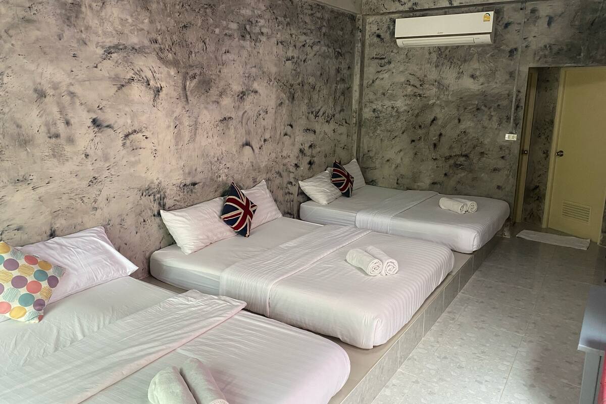 Ein Zimmer mit drei Betten, jedes mit weißer Bettwäsche und Kissen, darunter ein Bett mit Union-Jack-Motiv. Es gibt eine Wand-Klimaanlage und eine Tür rechts.