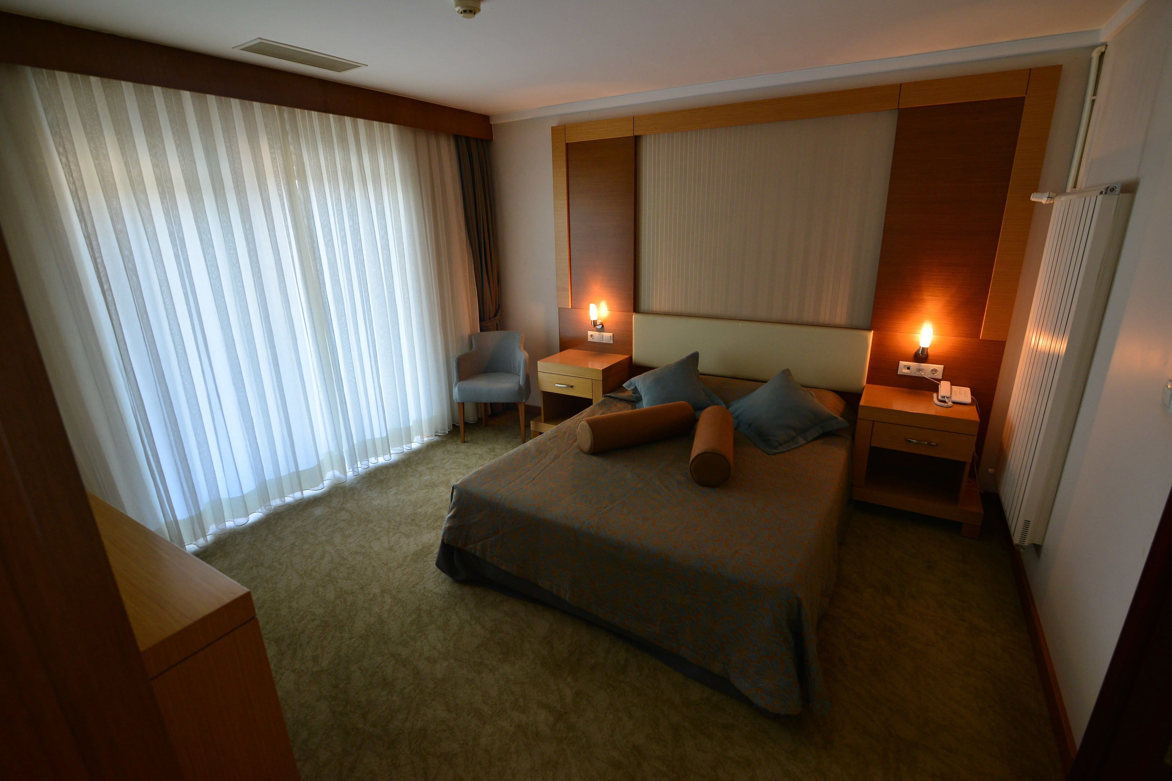 Quarto de hotel com uma cama grande, duas mesas de cabeceira, uma cadeira e uma janela com cortinas.