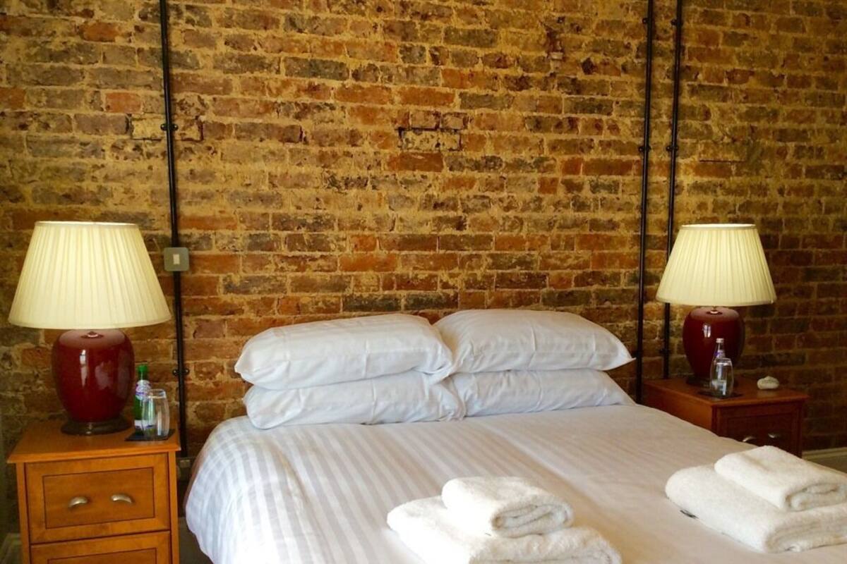 A neatly made bed with white linens and two pillows, a wooden nightstand with a lamp, and a brick wall in the background.