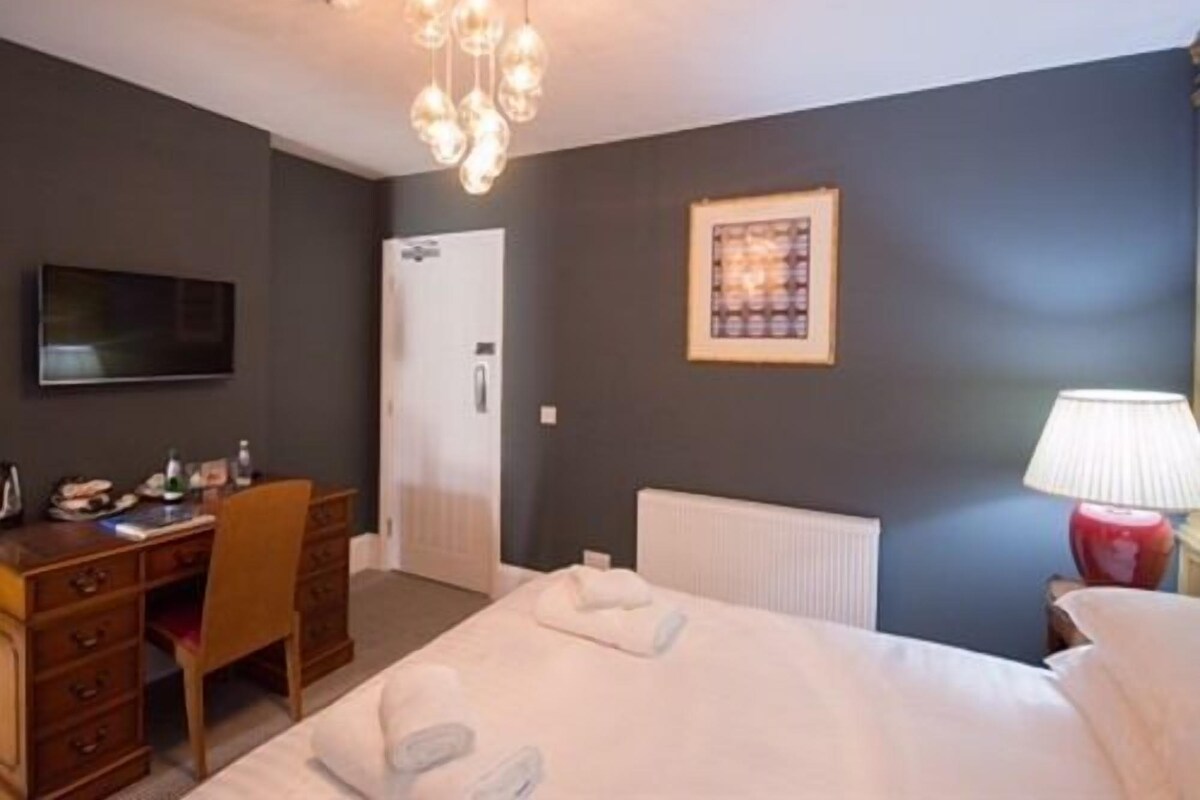 Quarto de hotel com cama, escrivaninha, cadeira, televisão, quadro emoldurado e abajur.