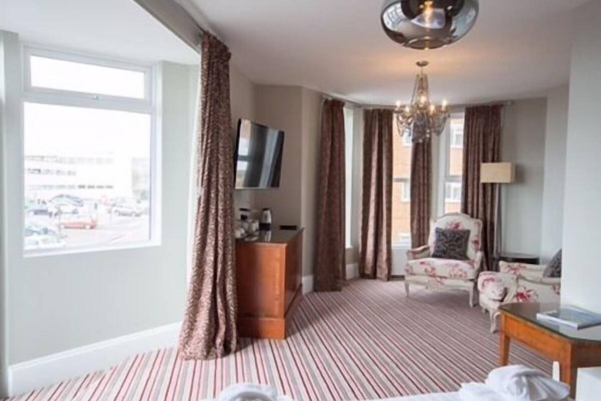 A hotel room with a striped carpet, a bed, a desk with a lamp, a chair, a television, and a window with curtains.