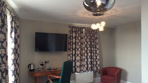 A room with a flat-screen TV, a desk with a chair, a radiator, patterned curtains, and a hanging light fixture.