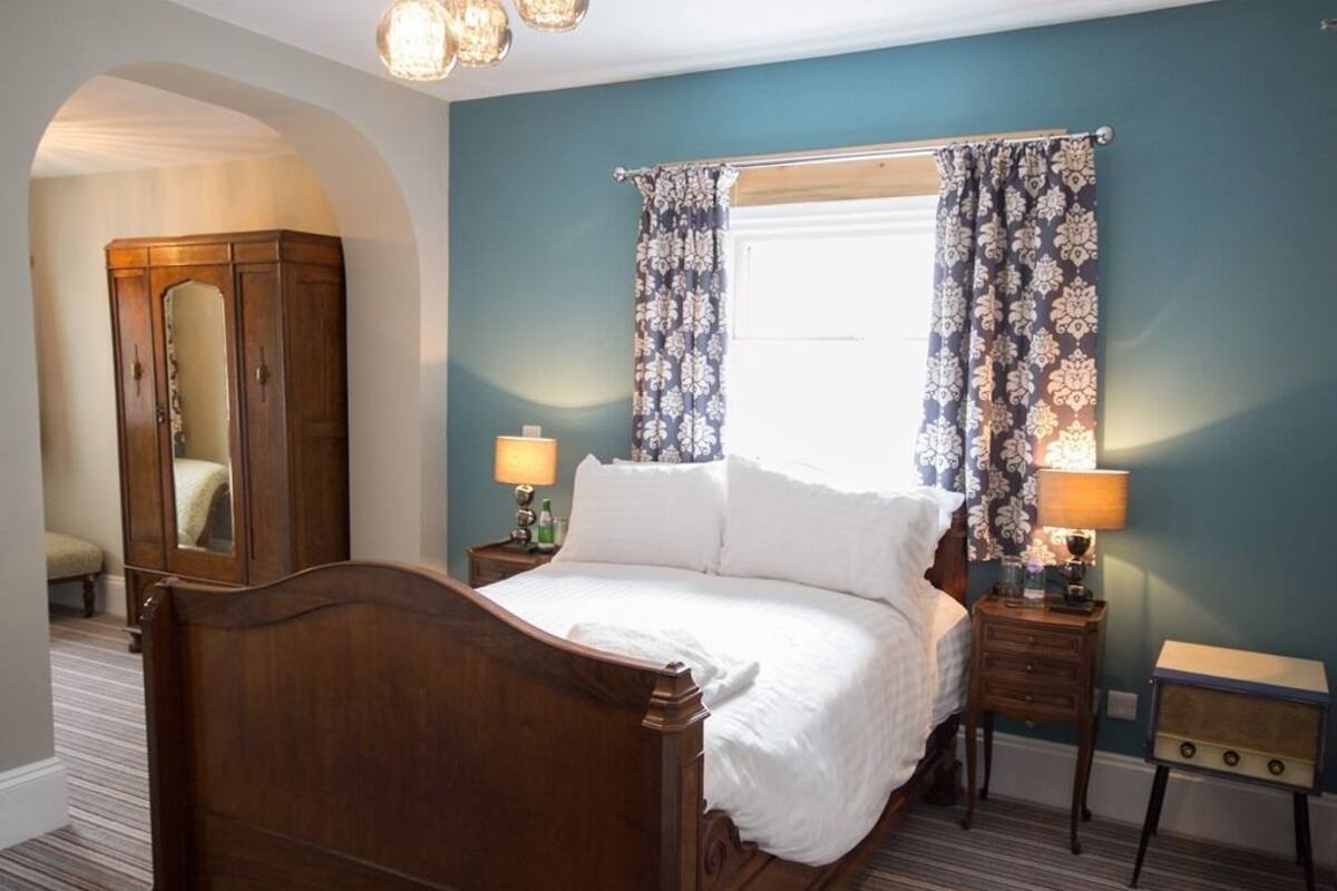 A bedroom with a wooden bed, two bedside tables with lamps, a wardrobe, and a window with patterned curtains.