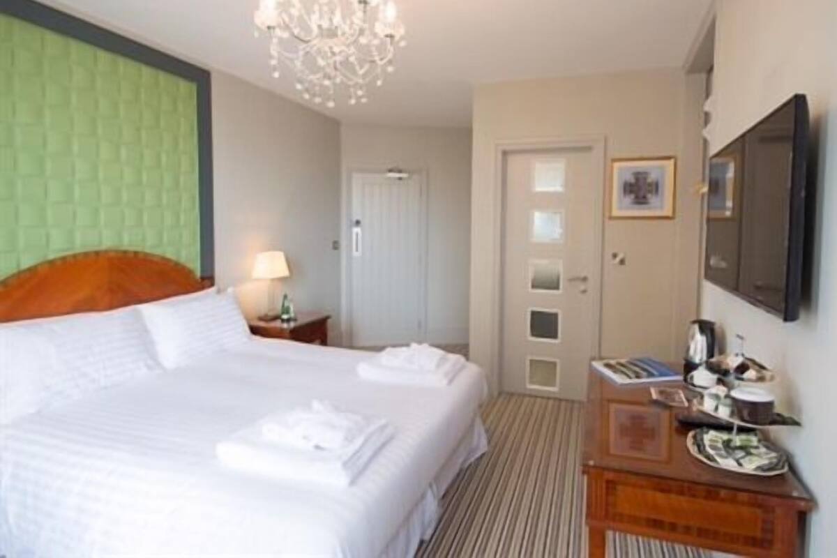 A hotel room with a large bed, a chandelier, a green accent wall, a door with a small window, and a wooden dresser with a tray of items.
