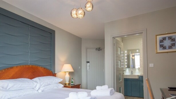 A hotel room with a large bed, a nightstand with a lamp, a bathroom visible through an open door, and a small table with a chair.