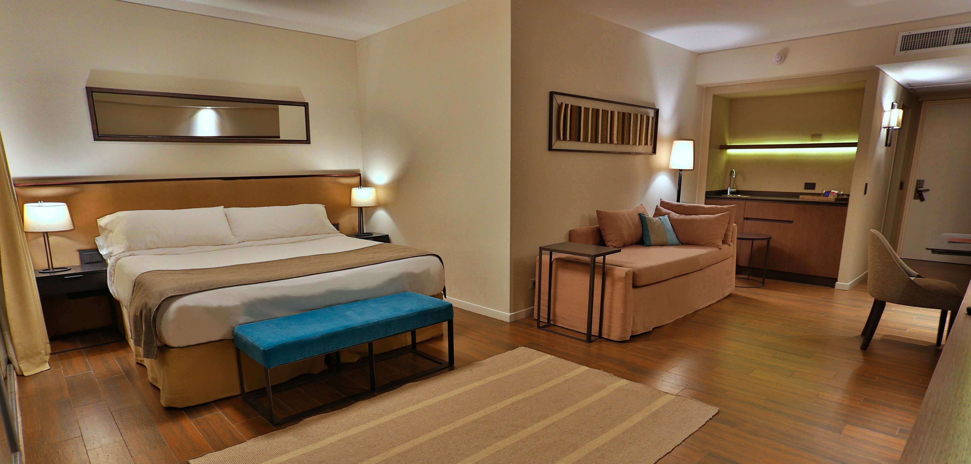 A hotel room with a bed, a bench, a sofa, a small table, and a kitchenette area.