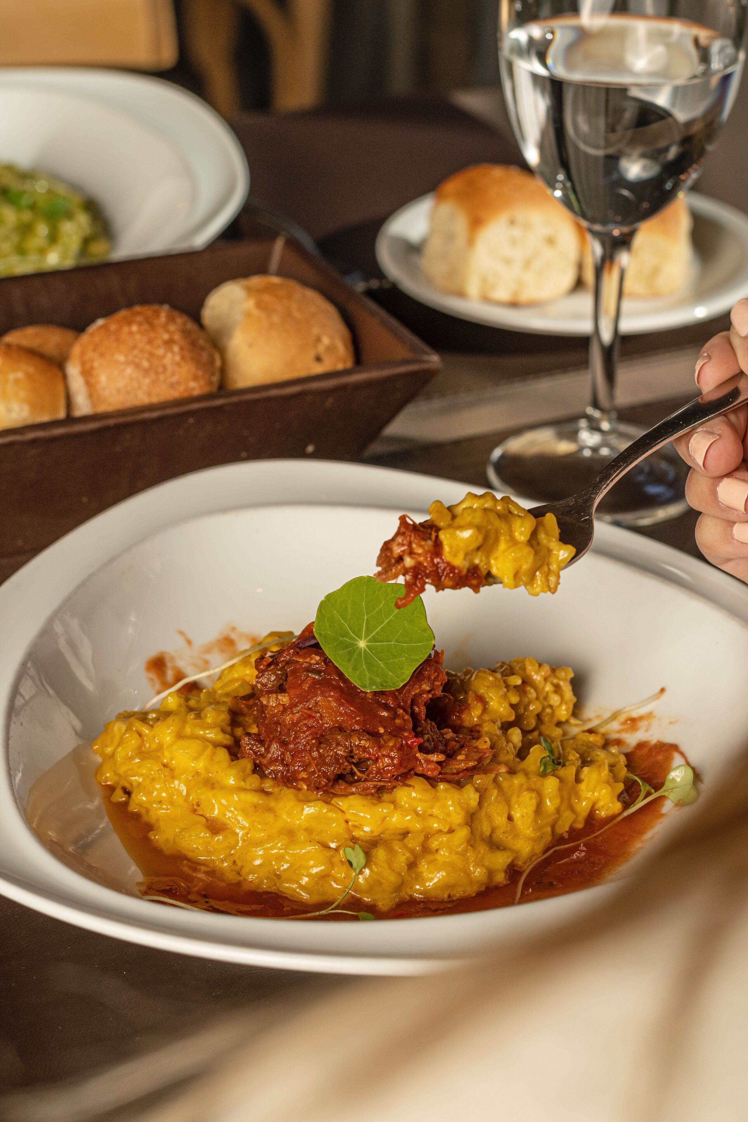 Una mesa de comedor con un plato de comida, una copa de vino y un bol de panecillos.
