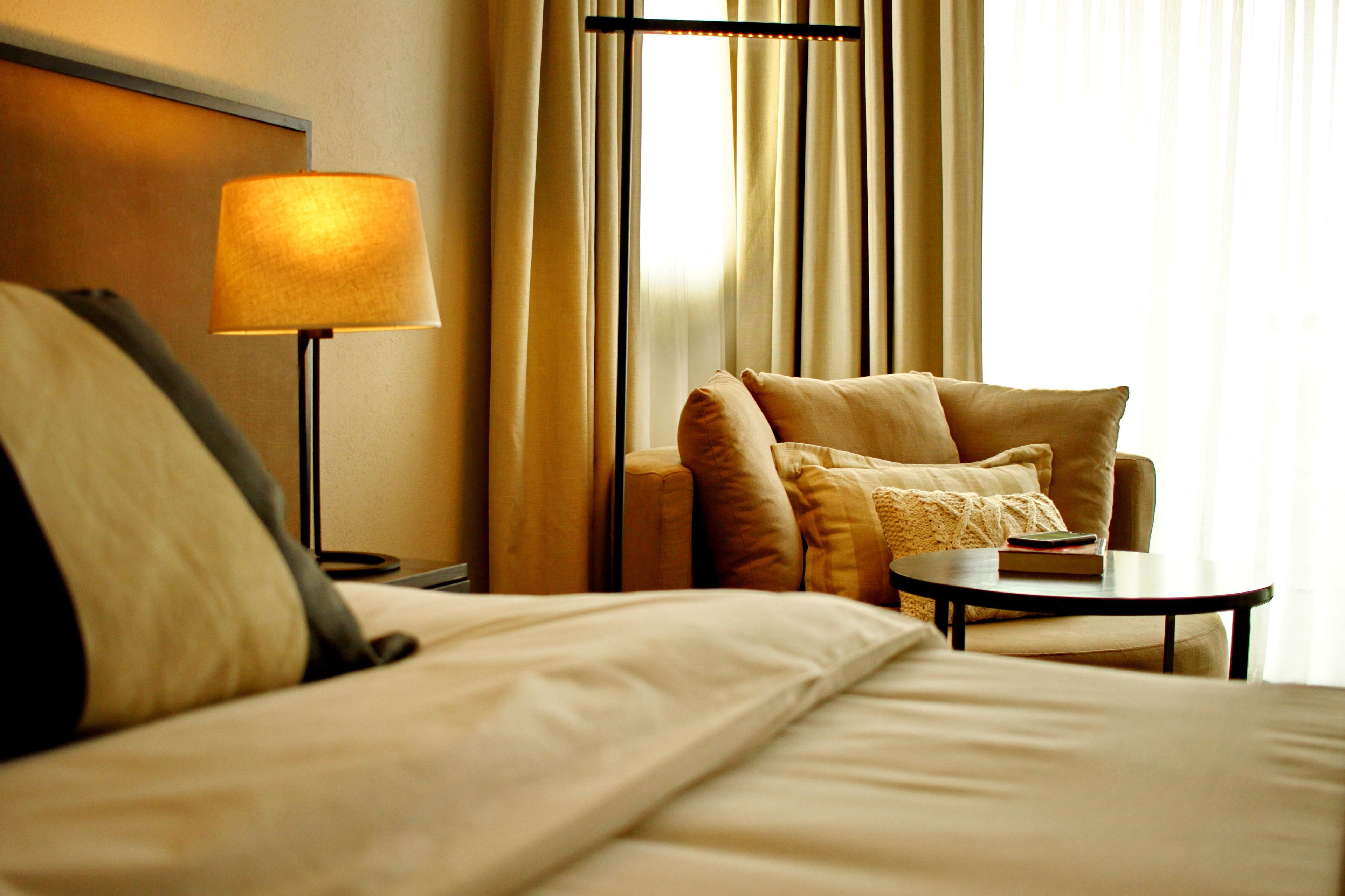 A hotel room with a bed, a chair, a table, a lamp, and curtains.