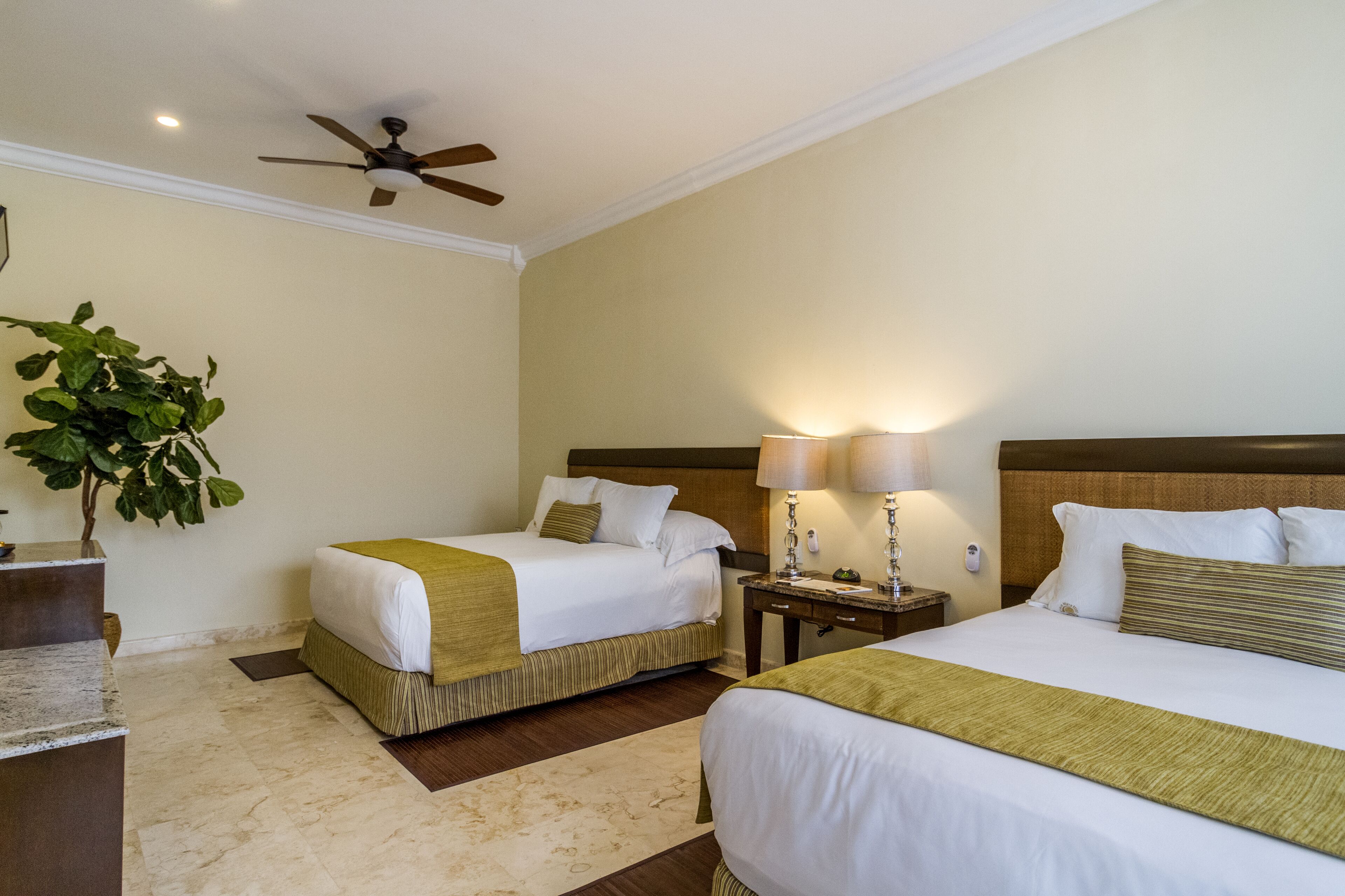 Une chambre d’hôtel avec deux lits, un ventilateur de plafond, une table de chevet avec une lampe et une plante en pot.