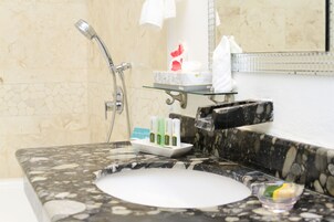 Un baño con lavamanos de mármol, cabezal de ducha manual y estante de vidrio con artículos de aseo.
