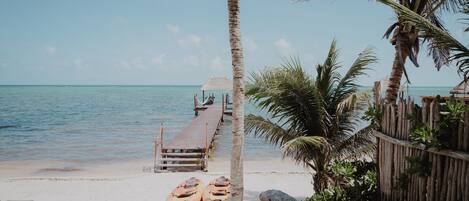 A beach with a wooden pier leading to a thatched-roof hut, kayaks, and a palm tree.