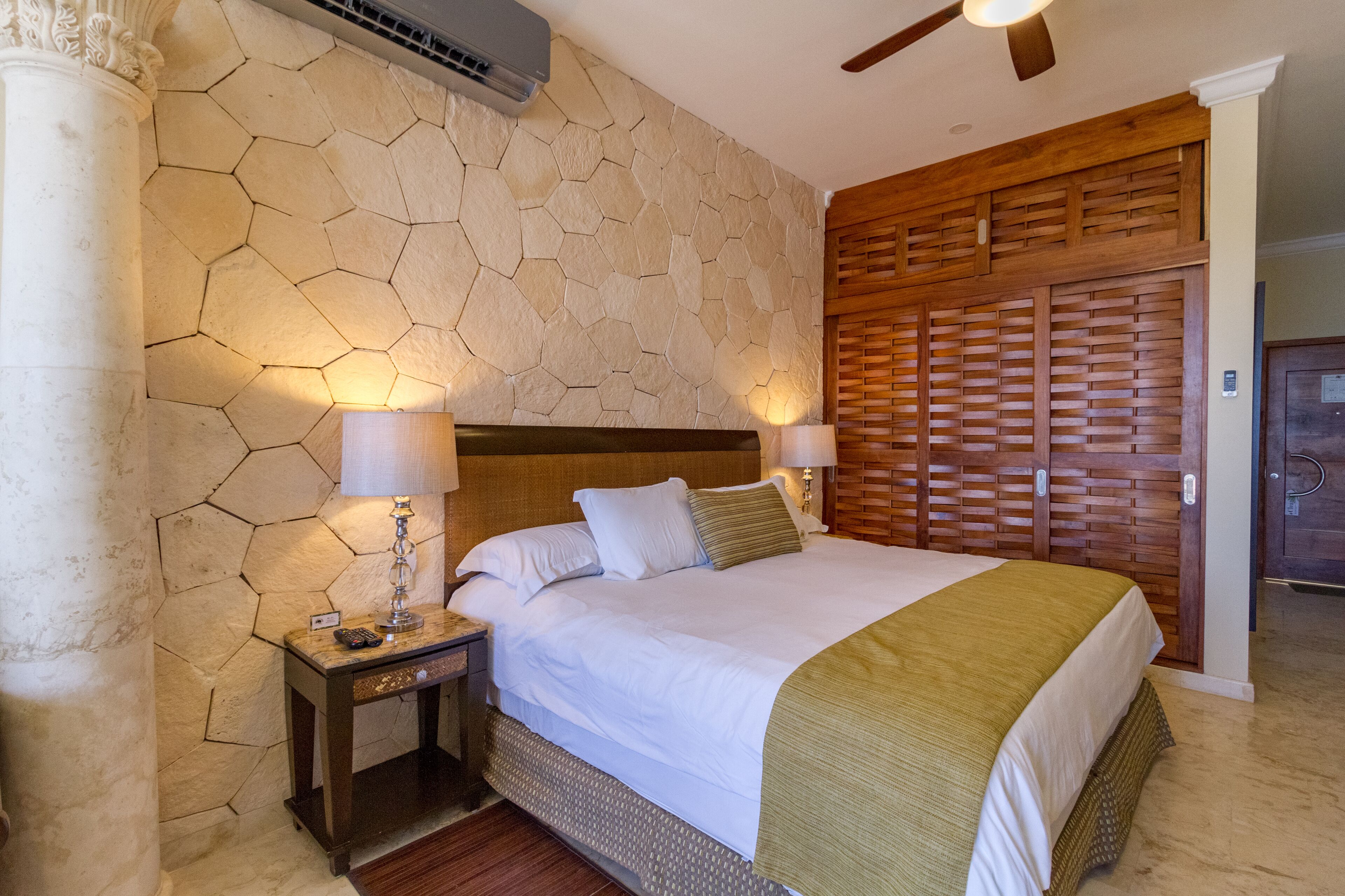 A hotel room with a stone-patterned wall, a wooden headboard, a bed with white linens, two bedside lamps, and a wooden wardrobe.