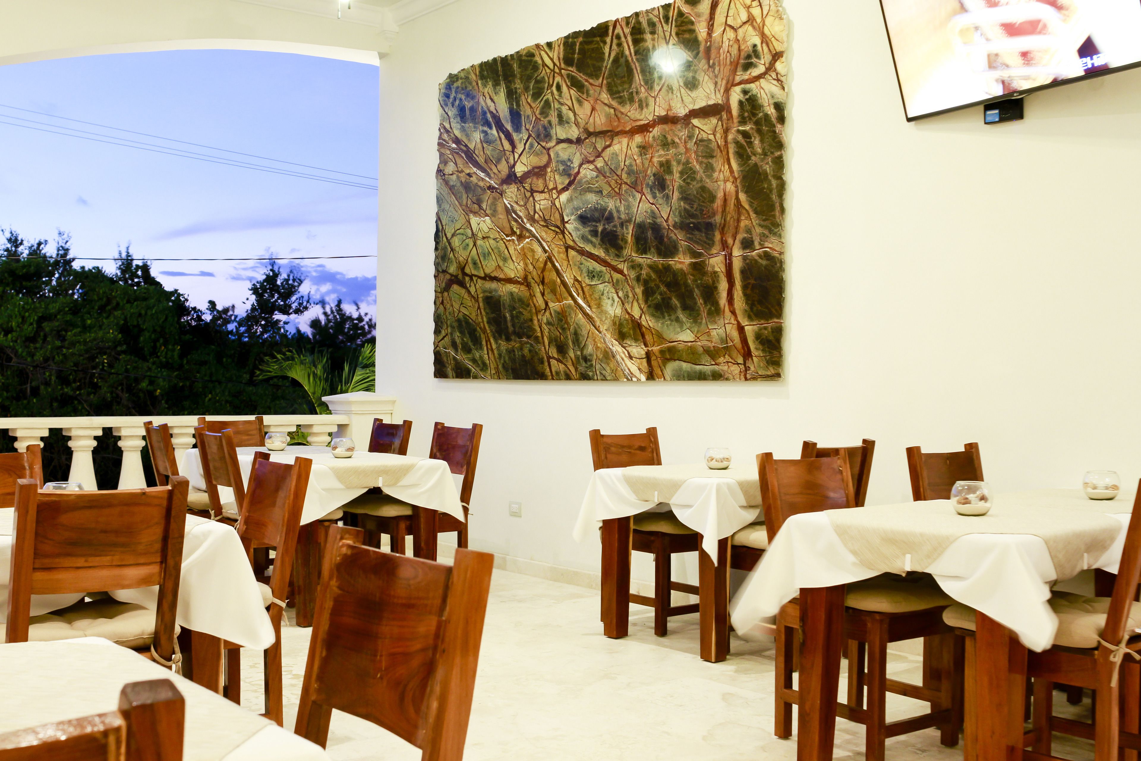 A dining area with multiple tables and chairs, a large abstract painting on the wall, and a view of trees and sky.