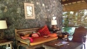 Um quarto com paredes de pedra, um sofá de madeira, uma mesa redonda com uma vela e flores, e uma cadeira.